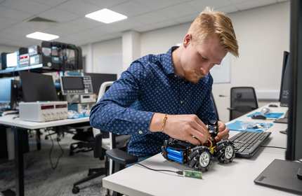 A person assembling a wheeled robot in an electronics lab, surrounded by tools and equipment.