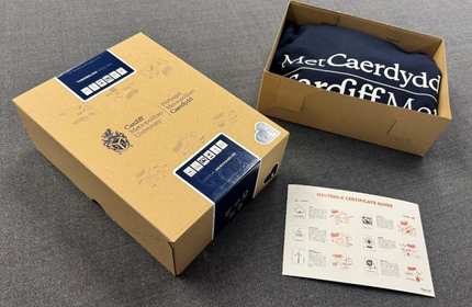 Two cardboards boxes, one opened and containing a Cardiff Met navy hoodie, on a carpeted floor