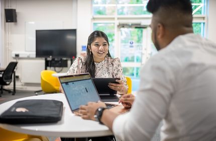 Two students working on laptops engage in discussion
