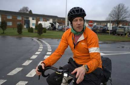 A man in high visibility clothing and a bicycle helmet rides a bicycle along campus roads