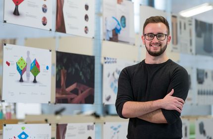 A young adult stands in front of a glass display board featuring pages of designs