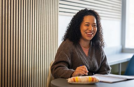A young woman sits and smiles at a table while working with a pen and pad of paper