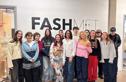 Group of fashion students and an instructor posing in front of a “FASHMET” wall.