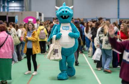 The Students' Union mascot, a light blue and white dragon, holding a tote bag in the middle of a busy Freshers' Fayre