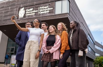 A group of six young adults taking a selfie in front of a building with the sign Cardiff Metropolitan University. They are smiling and appear to be enjoying their time together. The sky is partly cloudy.