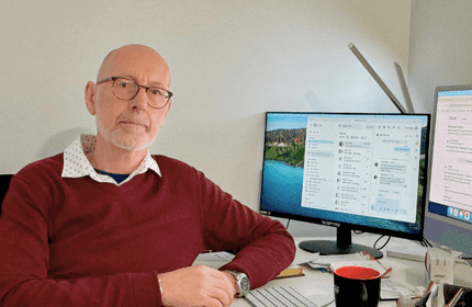 A bald man works at a desk with two computer monitors, engaged in tasks in a professional environment.