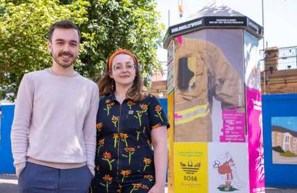A man and woman stand smiling outdoors next to a kiosk covered in colourful posters. The woman wears a floral jumpsuit and headband, and the man wears a beige sweater. Trees and a building are visible in the background.