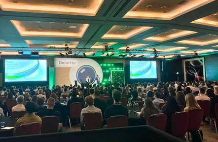 A large conference room filled with seated attendees facing a stage. The stage has a speaker, green lighting, and a screen displaying the Deloitte logo and presentation slides. The ceiling features recessed lighting.