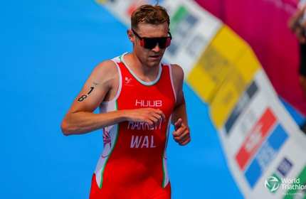 An athlete in a red triathlon suit is running with a blue track beneath. He wears sunglasses and has the number 18 on his left arm. The background shows colourful banners and the World Triathlon logo is visible at the bottom right.