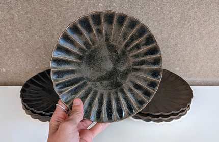 A hand holding a dark ceramic plate with a ribbed pattern. Several similar plates are stacked in the background on a white surface, against a neutral-coloured wall.