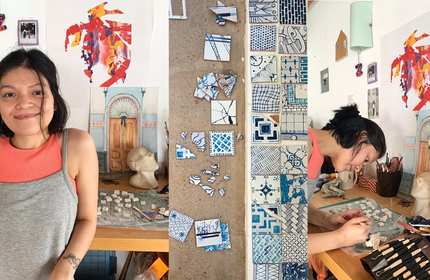 A woman stands and works at a table with mosaic pieces. Various small tiles are arranged on the table and a wall. Colourful artwork and a bust are visible in the background.