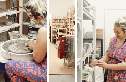 A woman shapes clay on a pottery wheel in a well-lit studio filled with pottery tools and materials.