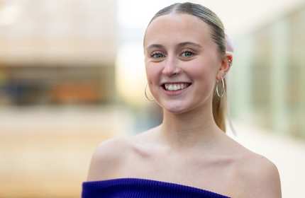 A young woman with hoop earrings and a nose ring smiles while wearing an off-the-shoulder blue top. The background is blurred, highlighting her cheerful expression.