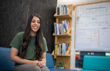 A woman with long hair sits on a blue sofa, smiling. She wears a green shirt and jeans. Behind her are bookshelves filled with books and a large poster on the wall. The setting appears to be a casual, modern office or study space.
