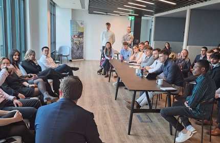 A group of people are seated in a modern conference room, attentively listening to a speaker at a meeting. The room has large windows and contemporary decor. Some attendees are taking notes, while others are engaged in the discussion.