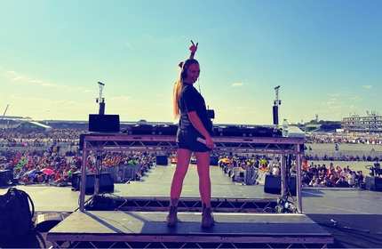 A DJ stands on an outdoor stage in front of a large, seated audience. The sky is clear and sunny. The DJ, with long hair, is making a peace sign with one hand. In the background, there are buildings and structures.