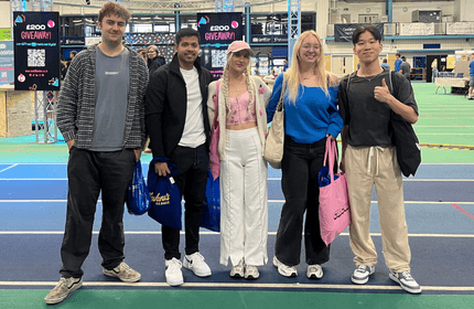 Five people standing together in a indoor athletic track, smiling and dressed in casual gear, enjoying their time at an organised event.