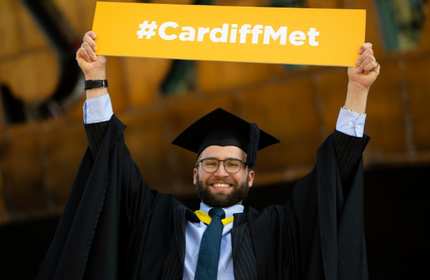 A graduate wearing a cap and gown joyfully holds up a yellow sign with #CardiffMet written on it.
