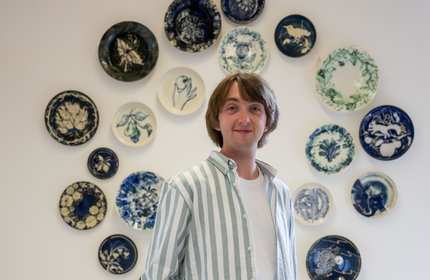 A person with shoulder-length hair stands smiling in front of a wall decorated with various blue and green patterned plates in circular arrangement. They are wearing a white and green striped shirt over a white top.