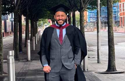 A Cardiff Met graduate stands between a row of trees wearing a grey suit and graduation cloak and cap