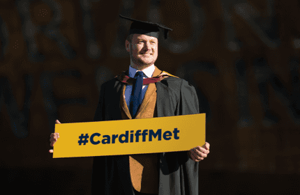 Ben Mason, wearing a graduation cap and gown, holds a large #CardiffMet placard