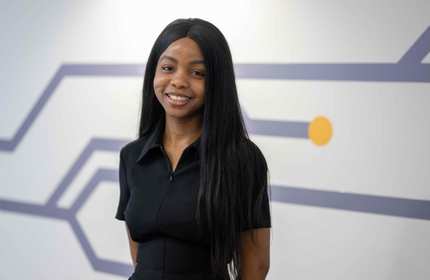 Elizabeth Aluko stands in front of a white wall with purple decorations