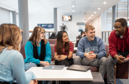 A group of give young people sit on a large sofa, engaged in discussion