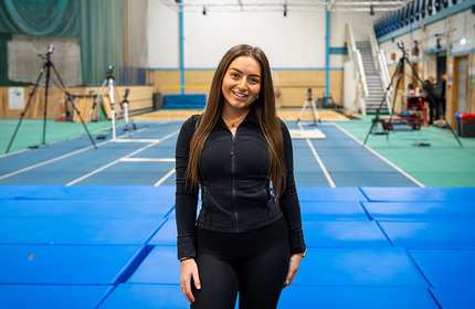 A young woman in black joggers and hoodie stands in the middle of a sports gymnasium