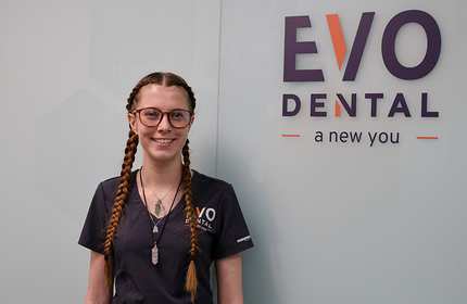 A young person with glasses and braided hair stands besides a wall branded with EvoDental