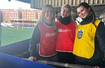 Three young women wear coloured sports bibs standing in the stands of a rugby stadium