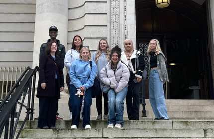 A group of young people stand at the top of a set of stairs outside the entrance of a large stone building
