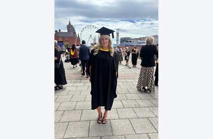 A Cardiff Met Graduate in cap and gown stands amongst crowds of fellow graduates and members of the public