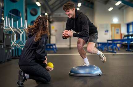 A young adult holding an orange ball with both hands balances on a board during exercise in a gymnasium