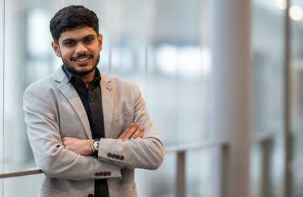 Jay Kiranbhai Patel, wearing a light gray suit and black shirt, stands in an open, airy building walkway