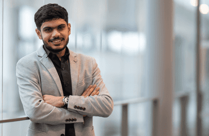 Jay Kiranbhai Patel, wearing a light gray suit and black shirt, stands in an open, airy building walkway