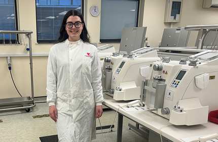 A young woman wearing a long white lab coat stands beside medical machinery in a laboratory