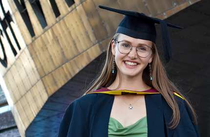 A Cardiff Met graduate, in cap and gown, stands underneath the iconic entrance of the Wales Millennium Centre