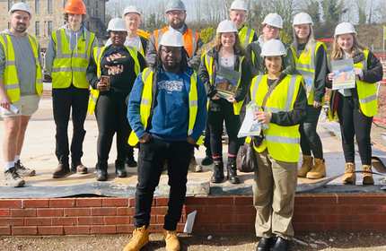 A large group of people wearing white safety helmets and high-visibility jackets stand on the foundations of a building on a construction site
