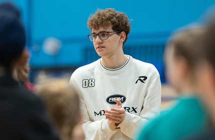 A young man with glasses and wearing a white sweatshirt clasps his hands together