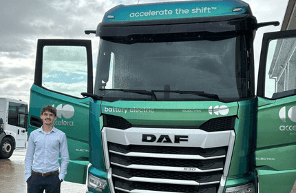 A young man in shirt and suit trousers stands in front of a large green lorry with both doors open
