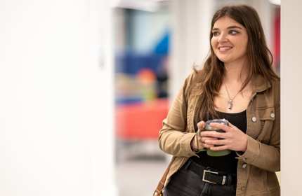 A young woman in a brown jacket holds a coffee mug with both hands while leaning against a wall