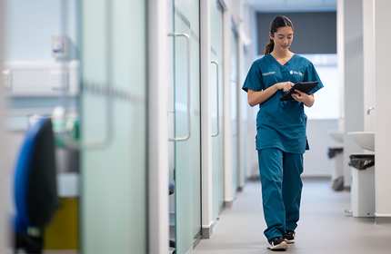 A young woman in blue-green medical uniform walks down a corridor working from an electronic tablet