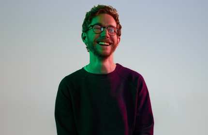 A young man wearing glasses smiles under low lighting