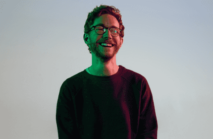 A young man wearing glasses smiles under low lighting