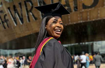 A young woman wearing a graduation cap and gown turns and smiles for the camera.