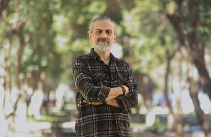 A mature student wearing a check shirt stands with arms folded on a large, sunny green space