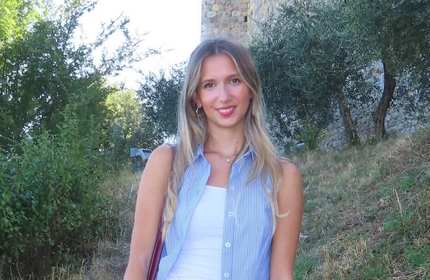 A young woman wearing a sleeveless blue shirt stands in front of some trees and an old stone building.