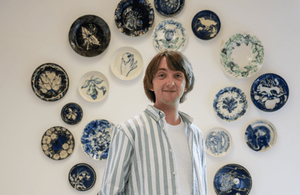 A person with shoulder-length hair stands smiling in front of a wall decorated with various blue and green patterned plates in circular arrangement. They are wearing a white and green striped shirt over a white top.
