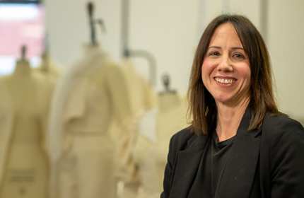 A woman in a black suit and shirt smiles at the camera. In the background are several tailor dummies.
