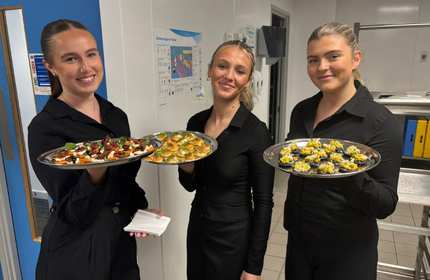 Three people stand in a professional kitchen holding trays of canapés.